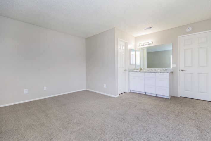 A spacious, empty room with light beige carpet and neutral-colored walls. On one side, there is a bathroom area featuring a double vanity with a mirror and light fixtures above. There is a door leading to the bathroom and a window allowing natural light to enter.