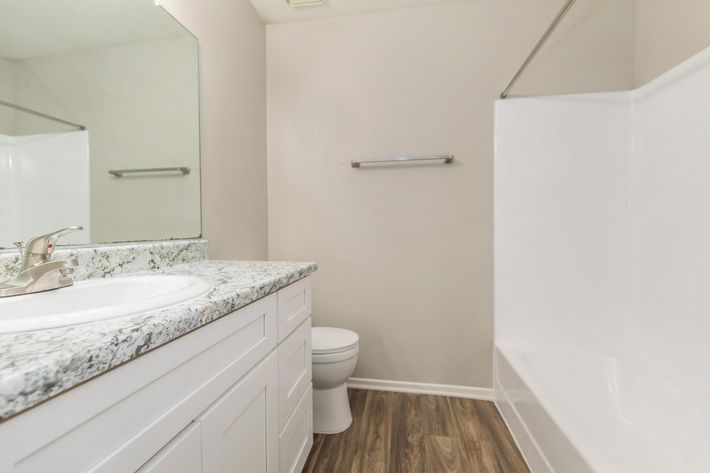 A modern bathroom featuring a white bathtub, a white toilet, and a marble countertop sink with a mirror above. The flooring is wooden, and there is a towel bar mounted on the wall. The walls are painted in a light color, creating a clean and spacious look.
