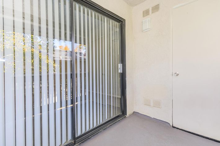 A balcony area featuring large glass sliding doors with vertical blinds, allowing natural light to enter. The floor is smooth and grey, and there is a plain door on the right side of the image. The walls are light-colored with simple textures.