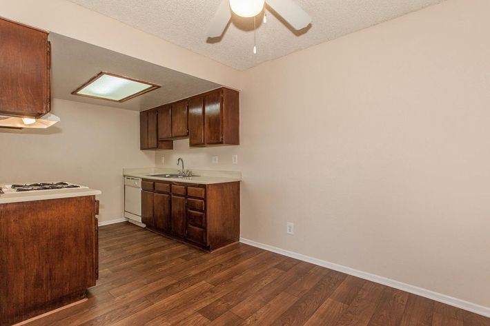 A spacious kitchen featuring dark wooden cabinets, a dishwasher, and a white countertop. The room has a ceiling fan and light-colored walls, while the floor is covered in light brown laminate. A small section of the ceiling has a fluorescent light fixture. Natural light is present, creating a warm atmosphere.