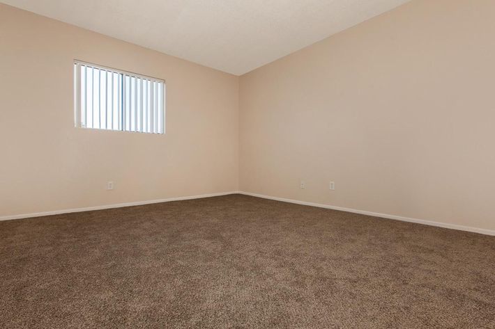 Empty room featuring beige walls and a carpeted floor. A small window with horizontal blinds allows natural light to enter. The space is devoid of furniture, creating a minimalist appearance. The overall atmosphere is neutral and spacious.