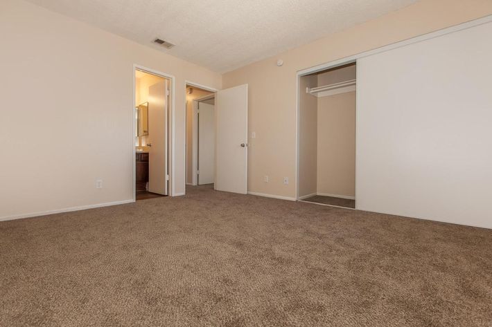 Empty room with beige walls and carpeted flooring. There are two doorways leading to a bathroom and a closet. The closet features sliding white doors, while the bathroom appears to be visible through the first doorway. The overall ambiance is neutral and spacious.