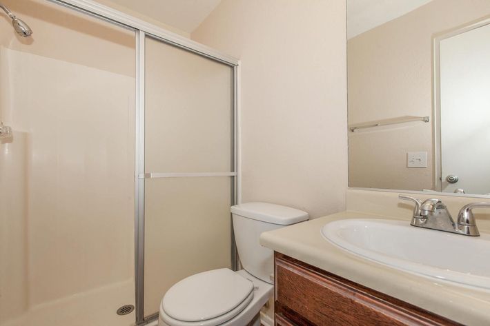 A small bathroom featuring a shower stall with a glass door, a white toilet, and a sink with wooden cabinetry. The walls are painted light beige, and there are minimal accessories visible, creating a clean and simple aesthetic.