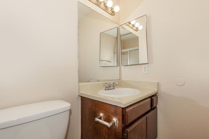 A small bathroom featuring a beige wall and a brown wooden vanity with a white sink. Above the sink are two mirrored cabinets and light fixtures. A toilet is positioned to the left of the sink, with neutral-colored flooring that complements the overall decor.