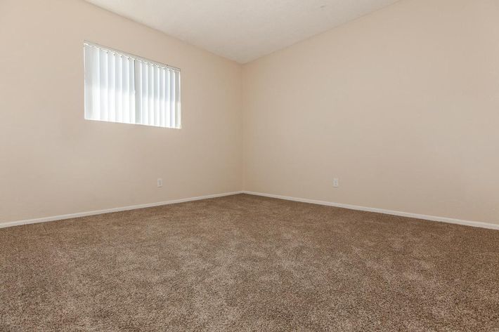 Empty room with light beige walls and a tan carpeted floor. A small window with vertical blinds allows natural light into the space. The room is unfurnished and has minimal decor, creating a simple and clean atmosphere.
