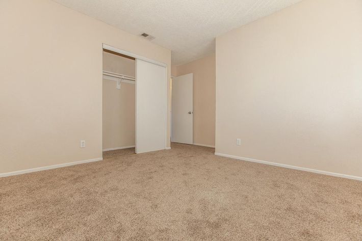 An empty room with beige walls and carpeted flooring. There is a closet with sliding doors on one side and a closed door leading to another area on the opposite side. The room is well-lit and spacious, lacking furniture or decorations.