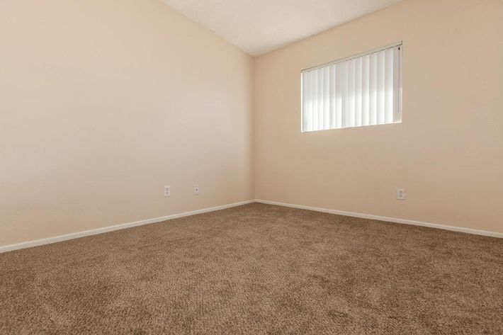 A vacant room with beige walls and a light brown carpet. There is one window with vertical blinds allowing some natural light inside. The space is empty, featuring two electrical outlets and minimal decor.