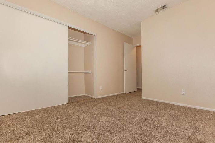 A spacious, empty room with light beige walls and carpet. There are two doors: one leads to a closet with sliding doors, and the other is an open door leading to another room. The overall atmosphere is bright and clean, suitable for various uses.