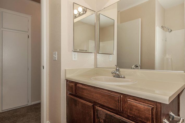 A clean bathroom featuring a light-colored countertop with a sink, wooden cabinetry, and two mirrors above the sink. A partially visible shower area is in the background, along with a door leading to a closet or another room. The walls are painted in neutral tones.