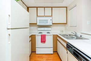 A modern kitchen featuring white appliances, including a refrigerator and a microwave. The stove is white with a red dish towel hanging on it. The cabinets are light-colored wood, and there are two sinks with a simple countertop. The floor is wooden, creating a warm and inviting atmosphere. Window treatments are simple blinds.