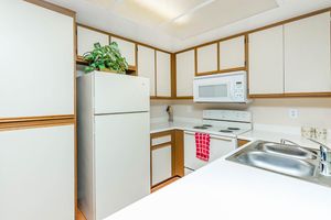A clean and modern kitchen featuring beige and brown cabinetry, a white refrigerator, a white microwave above the stove, and a double sink. A small potted plant is on the counter, adding a touch of greenery. A red dish towel hangs from the oven, enhancing the cozy atmosphere of the space.