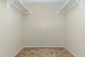 A blank, empty closet with beige walls and carpeted flooring. There are two white hanging rods on the walls for clothing, but no items are present in the space. The overall appearance is clean and minimalistic.