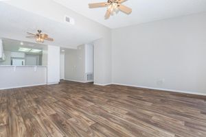 Spacious living area with hardwood-style floors, two ceiling fans, and neutral-colored walls. A kitchen is visible in the background, featuring a bar counter. The room is well-lit, creating a bright and open atmosphere.