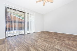 A spacious, unfurnished room with light-colored walls and a wooden floor. There are vertical blinds covering a sliding glass door that leads to an outdoor area. A ceiling fan is visible, and the lighting is bright, creating an airy atmosphere. The room is ready for personal decoration and furniture placement.