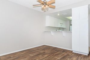 Interior view of a modern, unfurnished kitchen and living area. The space features light-colored walls, a ceiling fan with wooden blades, and a counter with a granite-like surface. The flooring is a warm wood tone, and the kitchen includes white cabinetry and appliances. Natural light brightens the area.