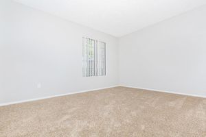 A spacious, empty room with light gray walls and a large window featuring vertical blinds. The floor is covered in beige carpeting, creating a soft and neutral atmosphere. The room is well-lit with natural light streaming through the window.