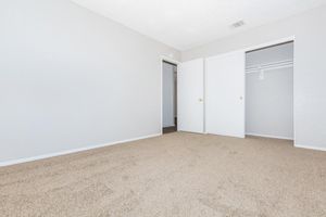 A spacious, empty bedroom featuring light gray walls, a beige carpeted floor, and two white closed doors leading to closets. The room is well-lit with natural light, highlighting its clean and simple design, making it suitable for various interior decorating styles.