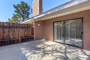 A patio area with a concrete floor, bordered by a wooden fence on one side and a wall on the other. Large sliding glass doors open into an indoor space, reflecting sunlight. The scene includes trees in the background, casting shadows on the patio surface.