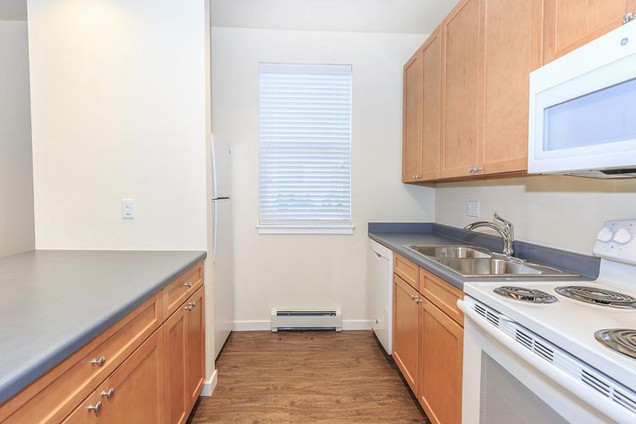 A modern kitchen featuring wooden cabinetry, a countertop, and stainless steel appliances. The space includes a stove, microwave, and dishwasher, with a refrigerator partially visible on the left. A window with blinds lets in natural light, and the floor is made of wood-like material.