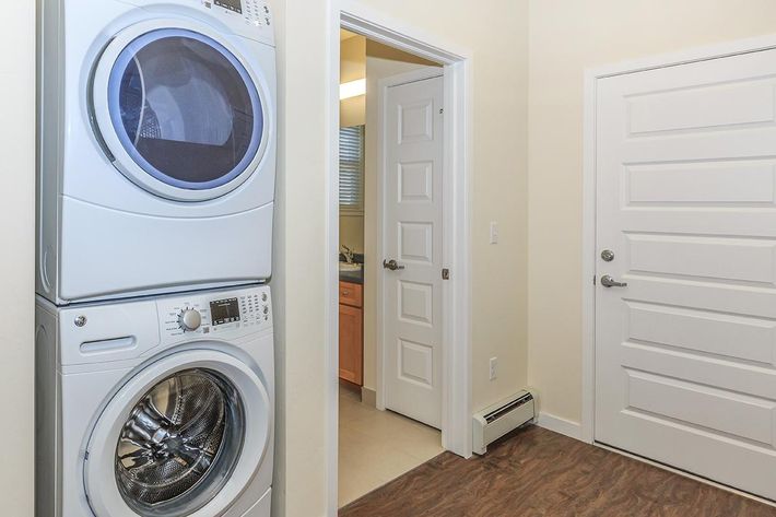 A laundry area featuring a stacked washer and dryer unit on the left, with a door leading to another room on the right. The walls are painted light colors, and the floor has a wood-like finish. A window provides natural light, and there is an air vent near the floor.