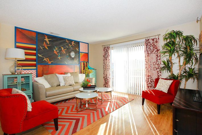 A bright and inviting living room featuring a large, colorful wall mural with birds, a beige sofa with decorative pillows, two red chairs, a glass coffee table, and a zigzag-patterned rug. Sunlight filters through sheer curtains, illuminating the space and a large green plant in the corner.