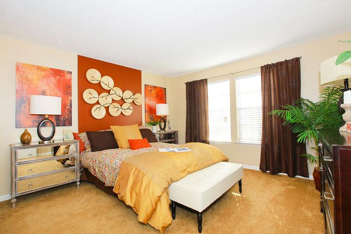 A cozy bedroom featuring a bed with colorful bedding, an upholstered bench at the foot, two stylish bedside tables with lamps, and abstract artwork on the walls. Large windows with brown curtains allow natural light to brighten the space, complemented by a potted plant for a touch of greenery.