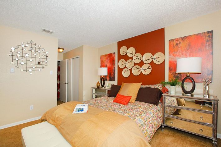 A stylish bedroom featuring a large bed with colorful floral bedding and orange accent pillows. The room has two decorative wall art pieces with a nature theme, mirrored nightstands, lamps, and a round wall mirror. Neutral-colored walls and tan carpet create a cozy atmosphere.
