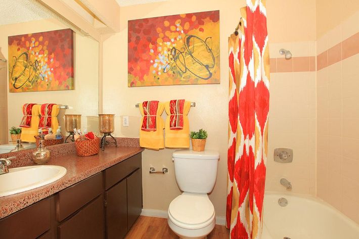 A bright and colorful bathroom featuring a sink area with a dark wooden cabinet, double sinks, and a large mirror. The wall displays an abstract painting with red, yellow, and orange hues. A vibrant red and white patterned shower curtain hangs beside a bathtub. Decorative towels and small plants add a cozy touch.