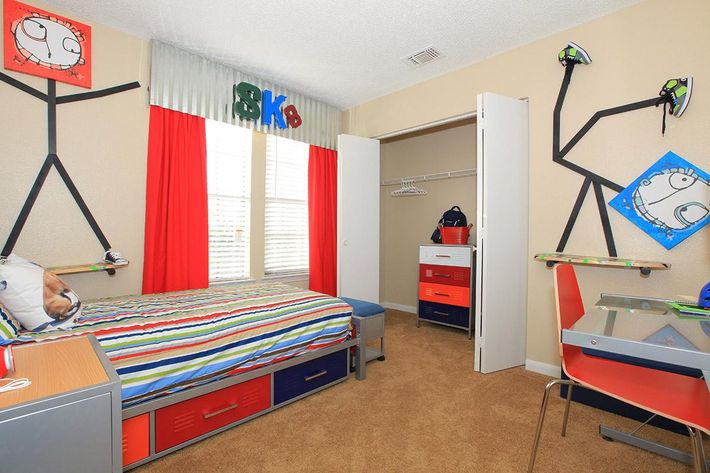 A colorful child's bedroom featuring a bed with a striped blanket, brightly colored curtains, and unique wall art. The walls are decorated with stick figure designs and shoe displays. There's a closet with a few clothes hanging, a modern desk, and drawers in primary colors. The carpet is a warm beige.