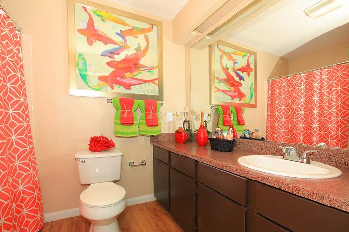 A bright and colorful bathroom featuring a large abstract painting on the wall, a white toilet, a double sink with a dark wood vanity, and an orange patterned shower curtain. Decorative items include a bouquet of flowers, red vases, and green towels, creating a vibrant and inviting atmosphere.