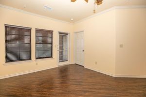 A well-lit empty room with light-colored walls and dark hardwood flooring. It features three windows with dark blinds, a door leading outside, and a closed door that likely leads to another room. The ceiling has a fan, and there's a subtle crown molding at the top of the walls.