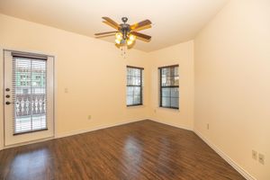 A bright, empty room with light brown walls and a wooden floor. It features a ceiling fan with lights, two windows with wooden blinds, and a door leading outside. The space is well-lit and has a clean, uncluttered appearance, ideal for various uses or decorating.