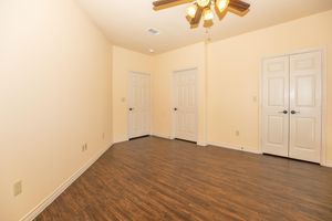 Empty room with light beige walls and a wooden floor. There are three closed doors—two on the right and one at the back. A ceiling fan with light fixtures is visible in the center of the ceiling. The room is well-lit and spacious, creating a simple, neutral environment.