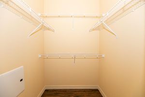 Empty closet with beige walls and wooden flooring. It features two sets of white wire shelves mounted on the walls, providing storage space. A small electrical panel is visible on the left side of the closet. The overall space is well-lit and appears neat and uncluttered.