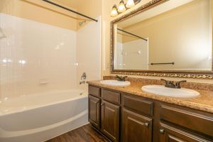 A well-lit bathroom featuring a white bathtub and shower combination, a large mirror above a double sink with dark wood cabinetry and a granite countertop. The walls are painted a light color, enhancing the space's brightness. Simple lighting fixtures are mounted above the mirror.