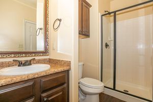 A bright and modern bathroom featuring a wooden vanity with a sink, a large mirror above it, and a glass-enclosed shower. There is a toilet beside the shower, and the walls are painted in a warm tone. The flooring has a dark wood appearance, adding a contemporary touch to the space.