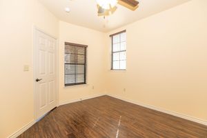 A vacant room featuring light yellow walls, a ceiling fan with wooden blades, and two windows with closed blinds. The floor is dark wood laminate, and there is a single door on one wall. The space appears well-lit, showcasing a minimalist and clean interior design.