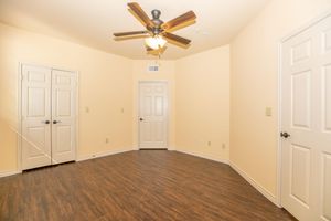 An empty room with light beige walls and dark wooden flooring. There are two closed white doors on opposite walls. A ceiling fan with wooden blades and a light fixture hangs from the ceiling, providing a bright atmosphere. The space is well lit and clean, suitable for various purposes.