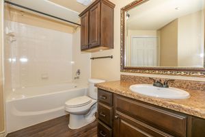 A clean and modern bathroom featuring a bathtub with a shower, a toilet, and a sink with a granite countertop. There is a dark wood cabinet above the sink, and a large decorative mirror is mounted on the wall. The walls are painted in a soft color, and the flooring is dark wood.