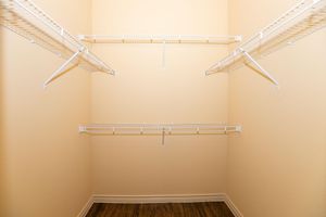 Empty closet with a light beige wall and wire shelving. The shelving consists of three horizontal racks positioned on the wall, designed for storage. The floor features a wood-like finish, creating a clean and minimalistic look. The space is well-lit and organized, ideal for storage or organizing items.