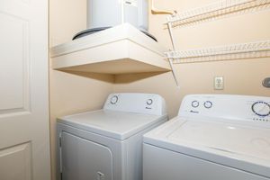 A typical laundry room featuring a stacked washer and dryer side by side. The appliances are white, and there is a shelf above for storage. The walls are painted a light color, and an electrical outlet is visible beside the dryer. A door is partially visible on the left side of the image.