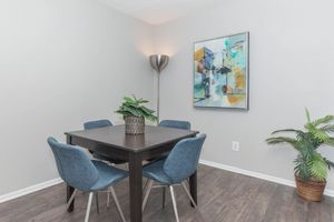 A modern dining area featuring a square black table surrounded by four blue upholstered chairs. A potted plant sits on the table, and a large abstract painting hangs on the wall. A floor lamp stands in the corner, and a potted plant is placed on the floor, adding color to the neutral-toned room.