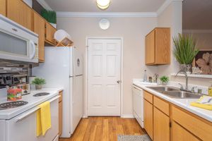 A clean and modern kitchen featuring wooden cabinets, white appliances including a microwave, refrigerator, and dishwasher. The countertop is equipped with a sink, and there are small plants and decorative items. A clock is mounted on the wall, and a door leads to another room. The floor is made of warm-colored wood.
