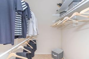 A neatly organized closet featuring various clothing items on hangers, including a striped shirt and a white blouse. The closet has a light-colored wall, a shelf with black and white storage boxes, and a decorative storage bin on the floor.