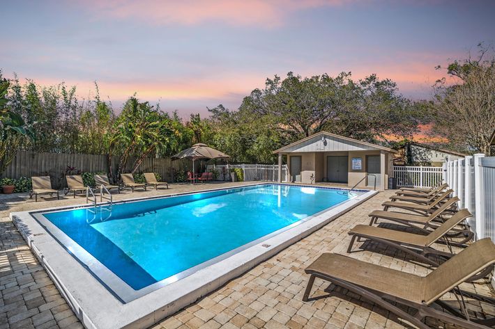A serene swimming pool surrounded by lounge chairs, set against a sunset sky. There are trees and greenery in the background, along with a small shed.