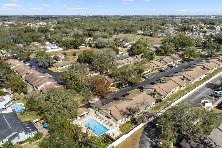 Aerial view of a suburban neighborhood featuring several single-story homes with brown roofs, a swimming pool, and extensive greenery in the background. The area includes parked cars along the streets and well-maintained landscaping.