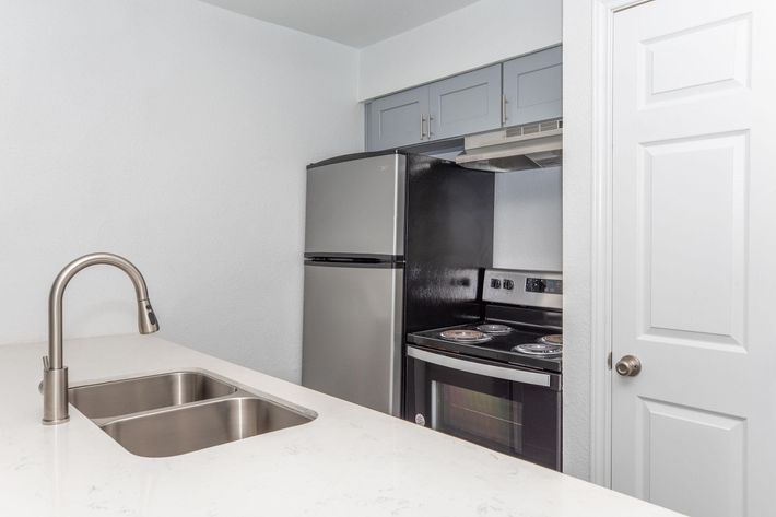 A modern kitchen featuring a stainless steel sink with a sleek faucet, a gray and black refrigerator, and an electric stove with an oven. The kitchen has light-colored walls and cabinetry, with a white door leading to another room.