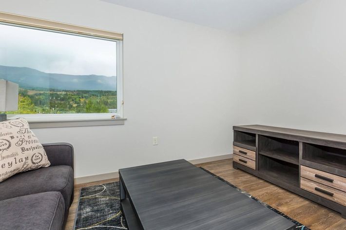 A minimalist living room featuring a dark gray sofa with decorative pillows, a low black coffee table, and a TV stand with drawers. There’s a large window revealing a scenic view of mountains and trees, with foggy weather outside. The walls are light-colored, enhancing the spacious feel of the room.