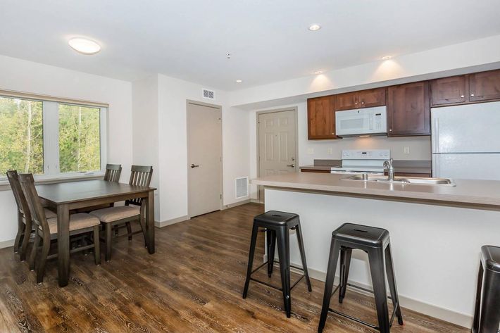 Bright and modern kitchen and dining area featuring a wooden table with four chairs, black bar stools, and sleek countertops. A white refrigerator and microwave are visible, along with natural light streaming in through a window, creating a welcoming atmosphere.