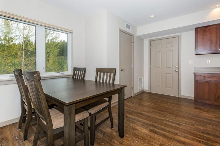 A modern dining area featuring a wooden table surrounded by four chairs. A large window offers a view of trees outside, while a doorway leads to another room. The walls are painted in a light color, and the flooring is dark wood. A kitchen area is visible with wooden cabinetry in the background.